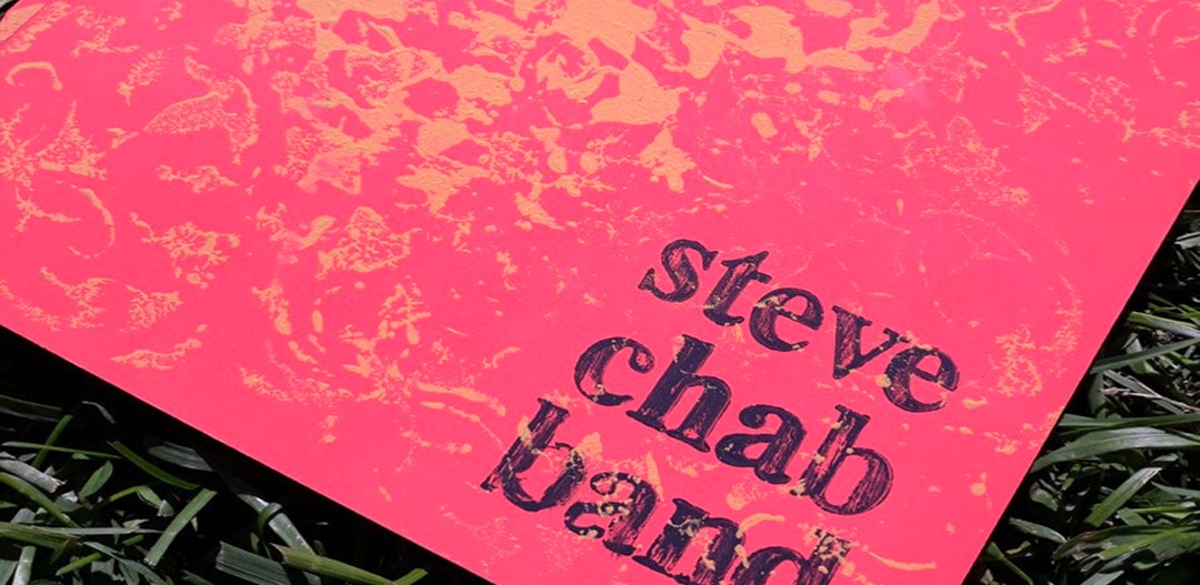 The corner of a hand-painted chapbook on grass. Gold paint stamped over salmon card, with steve chab band inked in dark type over the paint.